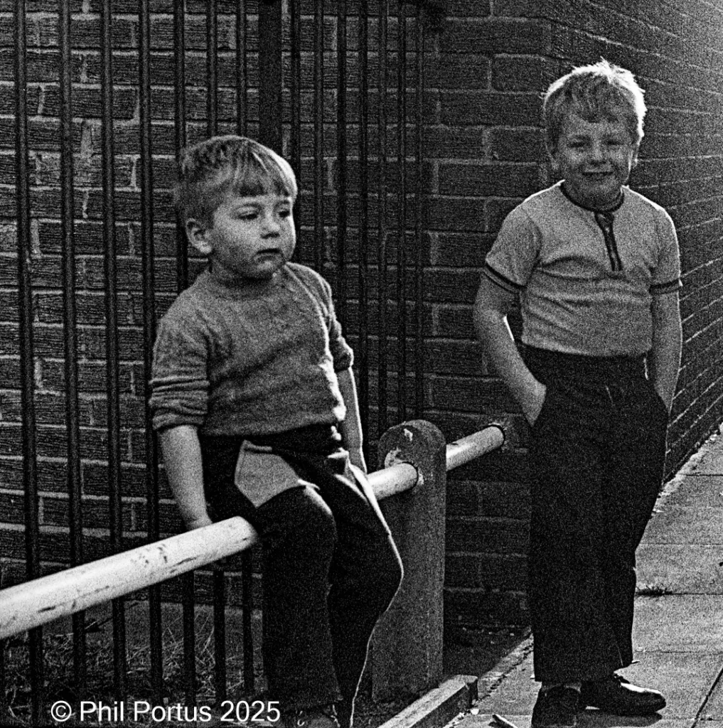 Two young boys sit and stand near a brick wall and metal railing in black and white.